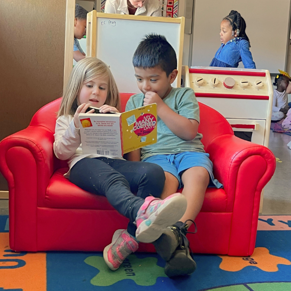  Two young children sit together on a small red chair reading the Dr. Seuss book "My Many Colored Days" in a preschool classroom.