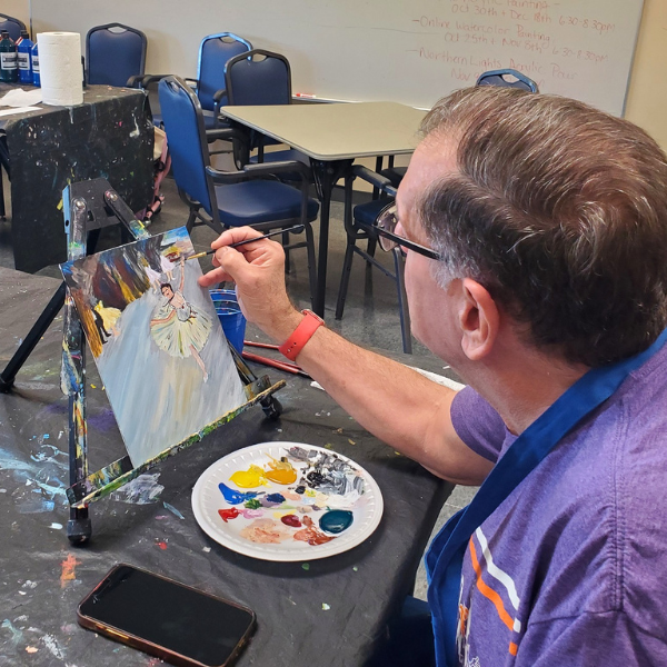  An adult man focuses on painting a ballerina on a small canvas during an enrichment class. He uses a fine brush and a paper plate as a palette for his paints.