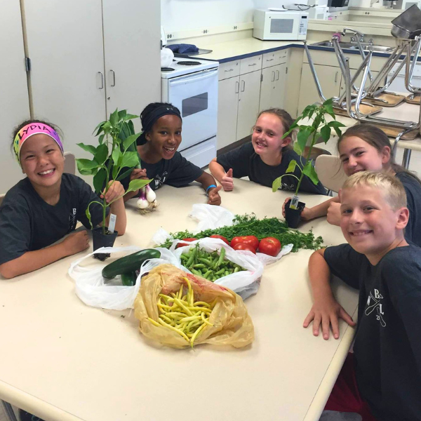  Five smiling students sit around a table in a classroom kitchen displaying a variety of fresh produce, including tomatoes, peppers, beans, and potted plants.