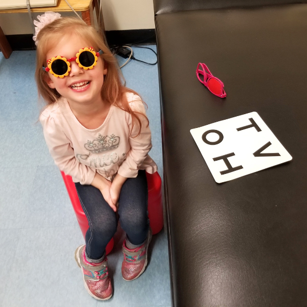 A young girl sits on a small red chair, smiling and wearing flower-shaped sunglasses. She is next to a black table with a few large letter cards and a pair of pink toy glasses on top.