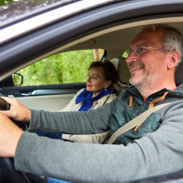 An older man smiles while driving a car with a woman in the passenger seat, both wearing seatbelts.