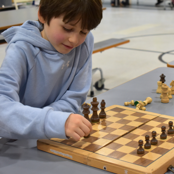  A young boy in a light blue hoodie focuses on a chess game, reaching out to move a dark chess piece on a wooden board.