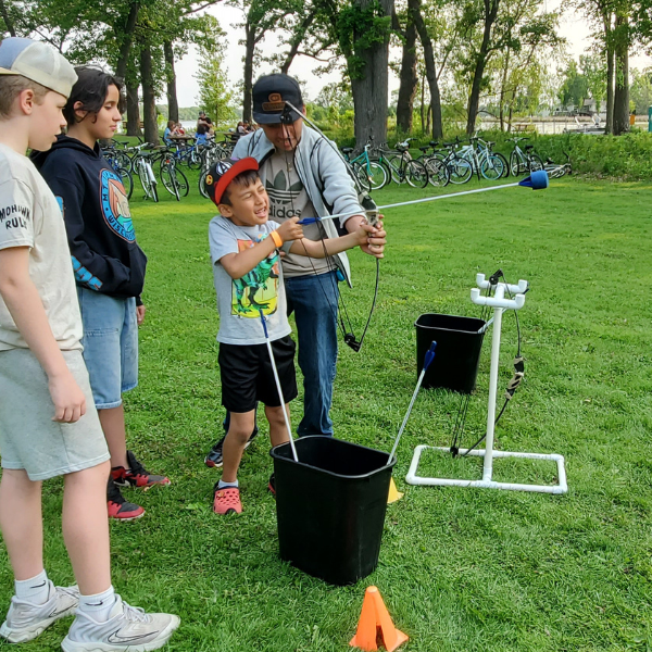 A father helps a young boy aim a bow and arrow during an outdoor archery lesson, while two other children watch. A row of bicycles and a lake are visible in the background.