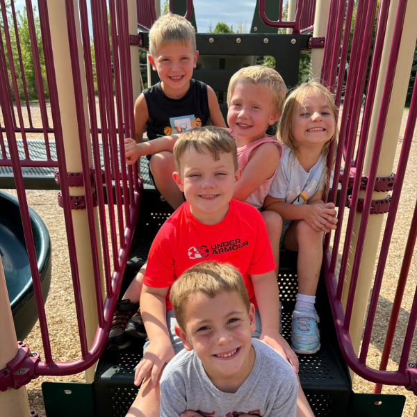 Five young children sit closely together on playground steps, smiling at the camera, framed by red metal railings on a sunny day.