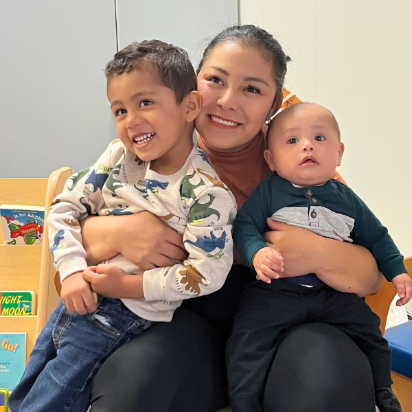 A smiling woman holds a happy young boy and a baby in her lap.