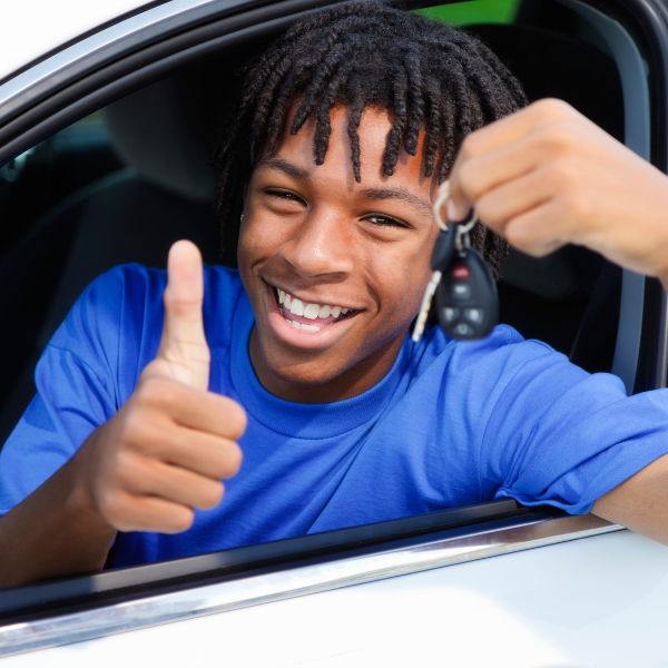 A smiling teen sits in the driver's seat of a car, giving a thumbs up and holding up a set of car keys.