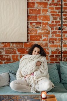 Woman relaxing wrapped in a knit blanket on a sofa against a brick wall.