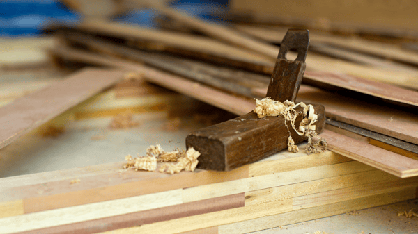 Hand plane woodworking tool resting on stacked wood boards with wood shavings scattered across a workshop table.
