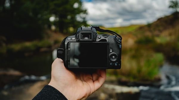 Close-up of a hand holding a camera in an outdoor setting with nature in the background.