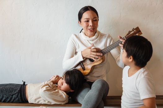 Cheerful slim female musician playing acoustic ukulele while sitting with crossed legs in apartment near charming sons in casual wear