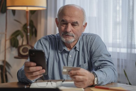 An elderly man using a smartphone and credit card for online shopping at home.