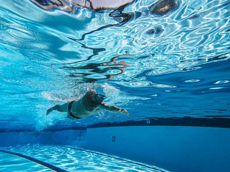Dynamic underwater photo of a man swimming freestyle in a pool wearing goggles and a cap.