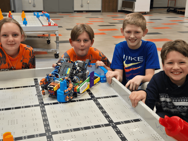 team of youth smiling while posing next to the VEX Robotics competition arena with robot they built