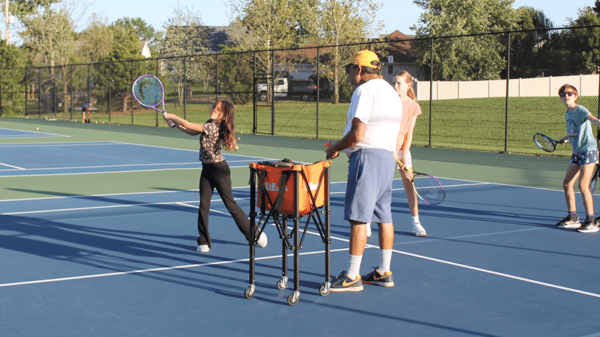 Tennis Coach Mark Platt standing next to a ball cart observing a student as they swing their tennis racquet at a practice ball.