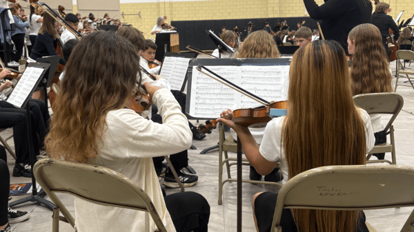 Two students seated in chairs facing toward their director while reading sheet music and playing their violins.