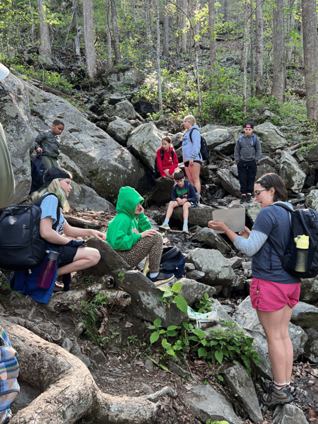 A wilderness instructor from the Great Smoky Mountain Institute at Tremont is presenting to a group of students who are seated on large rocks near tree roots.