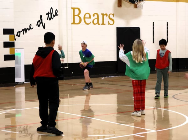 Students playing flag football in a gymnasium. The wall behind the students reads; "Home of the Bears."