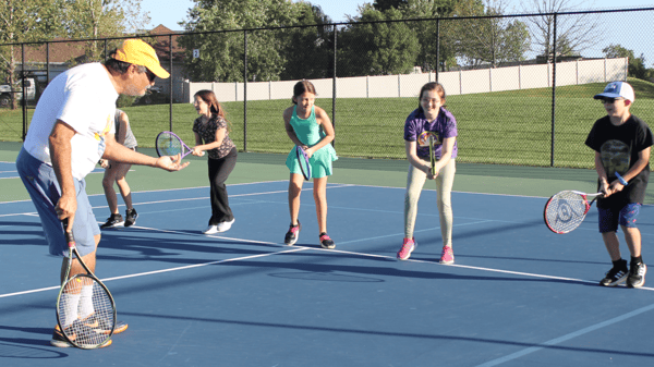 Coach Mark Platt with a tennis racquet in the foreground teaching a small group of middle school students also holding racquets on an outdoor tennis court.