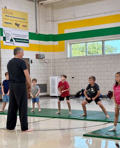 A martial arts instructor is facing a group of children practicing postures on mats in a gymnasium. 