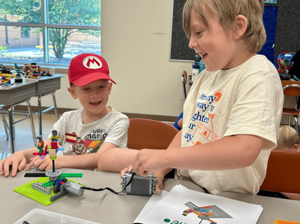 Two boys at a classroom table. One is operating a battery pack attached to a structure they have built with LEGO® bricks. The battery pack give power to rotors and gears attached to their design, allowing a mini figurine to spin on top of it.