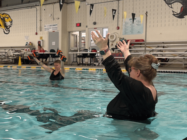 Two adults with arms raised while standing in chest deep water at an indoor pool. One is guiding the other through aquatic exercise.