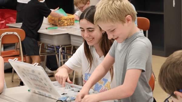 A student and Bricks 4 Kidz instructor are seated at a table with the lid raised on a supply kit. The student is building with LEGO® bricks.