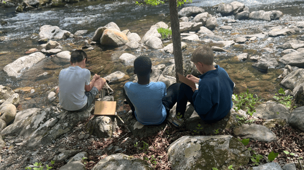 Three students sitting together on rocks by a creek at the Great Smoky Mountains Institute at Tremont. Two students are writing about their observations in notebooks.