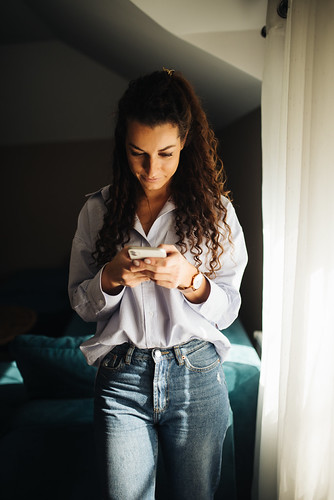 Woman with curly hair checks her smartphone while standing by a window in a cozy room during daylight hours