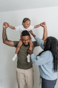 Father joyfully lifts his smiling child on shoulders, supported by mother indoors.