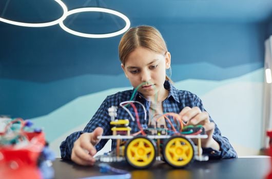 Teenage girl working on a robotic car project with wires and wheels inside a classroom.
