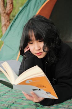 A young woman relaxing outdoors, reading a book near a camping tent.