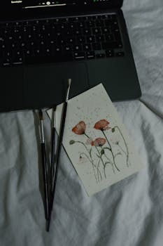 A creative workspace featuring a laptop, paintbrushes, and a floral watercolor painting.