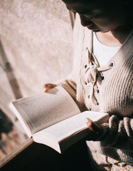 A young woman in a cozy sweater reading a book by a sunny window.