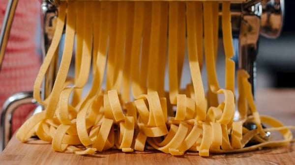 Close-up of fresh pasta strands being made with a pasta machine in an indoor kitchen setting.