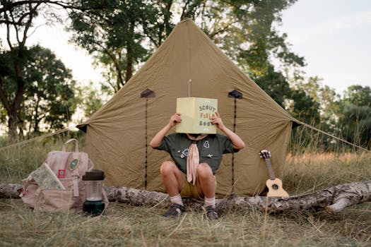 Young scout reading a field book in front of a tent, surrounded by camping gear and nature.