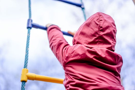 A child climbing a rope ladder at a playground, facing away, wearing a red jacket.