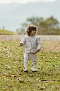 A joyful toddler walks on grass on a sunny autumn day, dressed warmly in cozy attire.