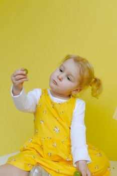 Cute toddler in a yellow dress thoughtfully holding grapes against a yellow background.