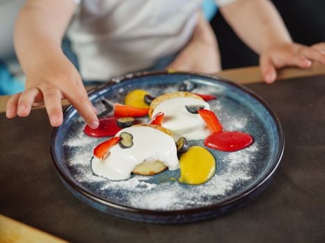 Close-up of a child's hand reaching for pancakes topped with fruit and sauce on a blue plate.
