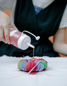 Unrecognizable master pouring paint from bottle on tied multicolored fabric placed on table with tablecloth while working using shibori technique