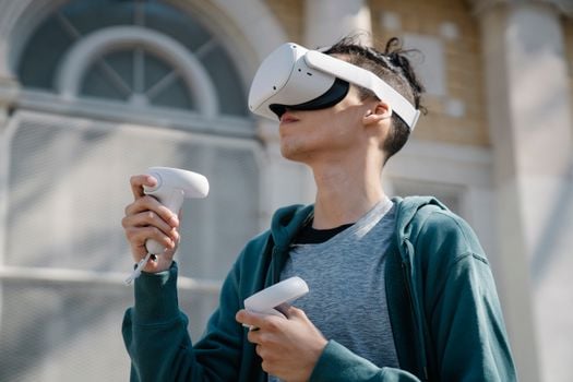 Low angle of young male in blue hoodie using modern wireless virtual reality headset and controllers near building on street