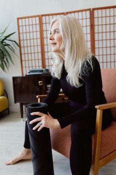 Senior woman in black long sleeves sitting with yoga mat, ready for exercise.