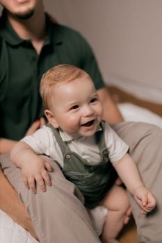 Smiling baby boy in green overalls sitting comfortably on a parent's lap indoors.