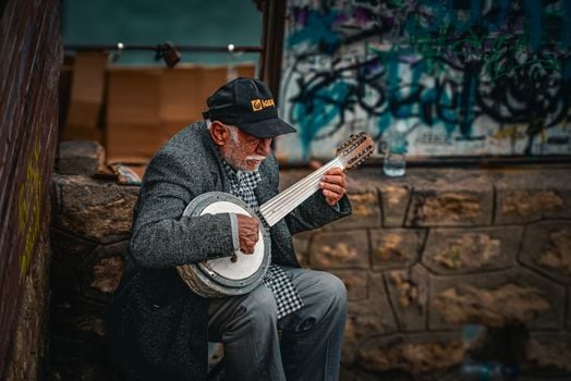 Elderly man in streetwear playing a banjo against graffiti wall. Urban music scene captured outdoors.