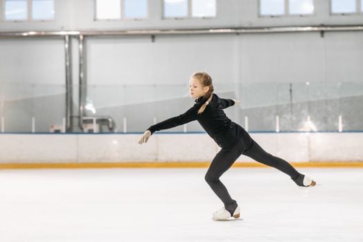 A young girl practicing figure skating moves at an indoor ice rink during the day.