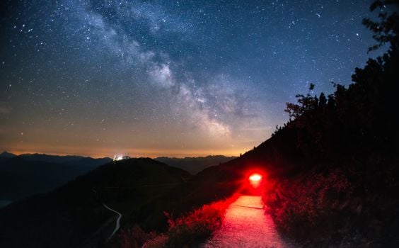 Nighttime view of the Milky Way over a mountain trail with red torchlight illumination.