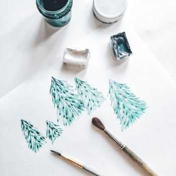 Top view of watercolor painting of Christmas trees with paintbrushes and paints.