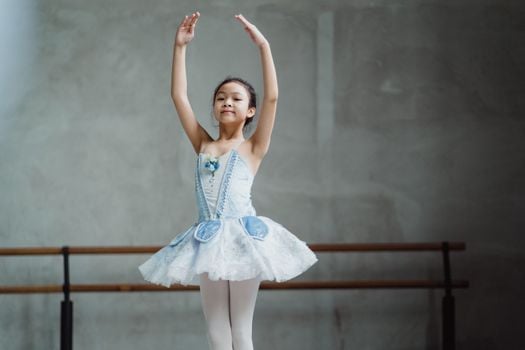 Little Asian girl dancing classical ballet with hands raised in studio equipped with barre