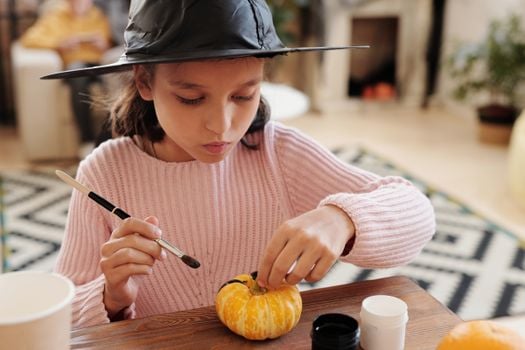 A young girl in a witch hat paints a small pumpkin indoors for Halloween fun.