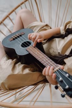 A young child playing the ukulele on a comfortable chair indoors.